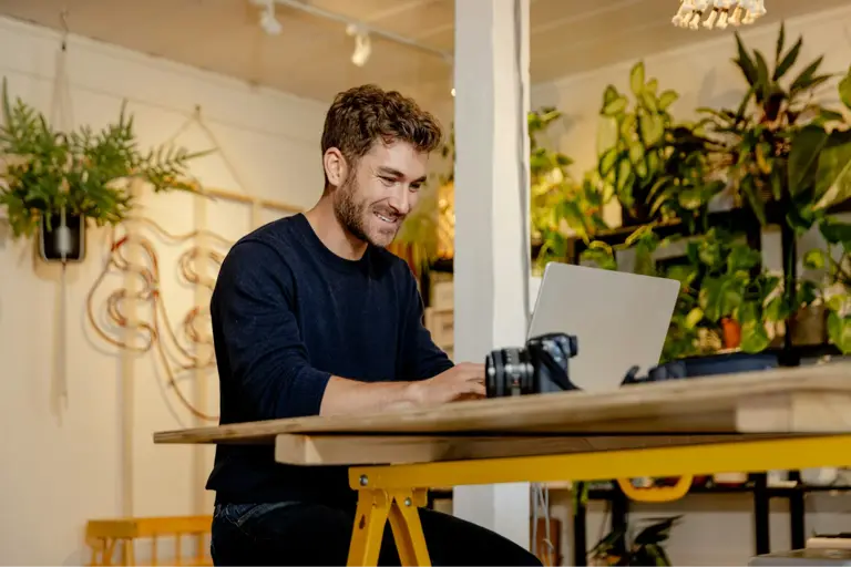 Mann an Schreibtisch mit Laptop, umgeben von grünen Pflanzen in hellem Gastronomieraum