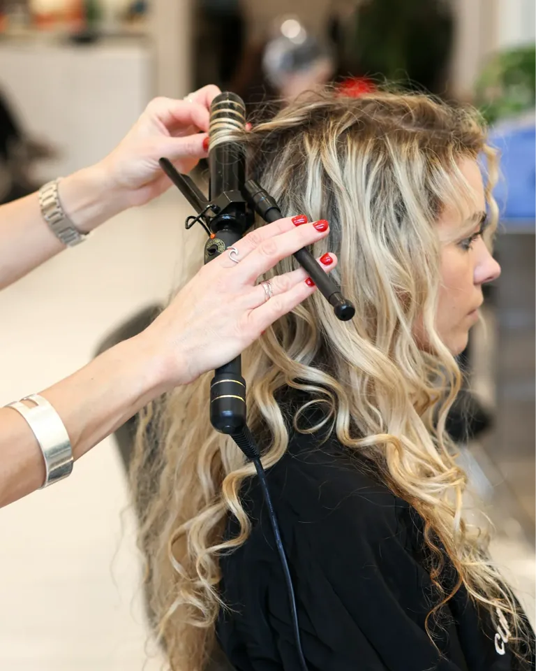 Friseur stylt blonde Locken mit einem Lockenstab in einem Salon.
