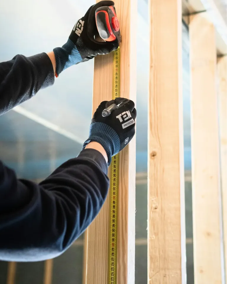 Handwerker in Arbeitshandschuhen misst Holzbalken mit Stahlmaßstab.