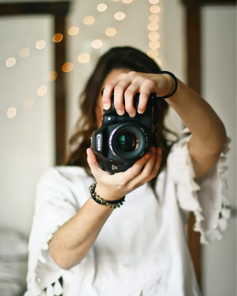 Person in weißem Shirt hält eine schwarze Digitalkamera vor das Gesicht, Bokeh-Lichter im Hintergrund.